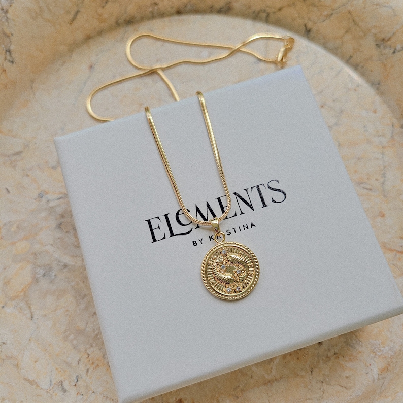 Puede incluir: Un collar de cadena dorada con un colgante redondo que presenta un s&iacute;mbolo del zod&iacute;aco y peque&ntilde;as piedras brillantes. El collar se muestra en una caja blanca con el texto "ELEMENTS BY KRISTINA".