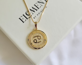 May include: Gold chain necklace with a round pendant featuring the Cancer zodiac sign. The pendant has a textured surface and a raised design of the zodiac symbol. The necklace is on a white background with the text "Elements by Kristina".