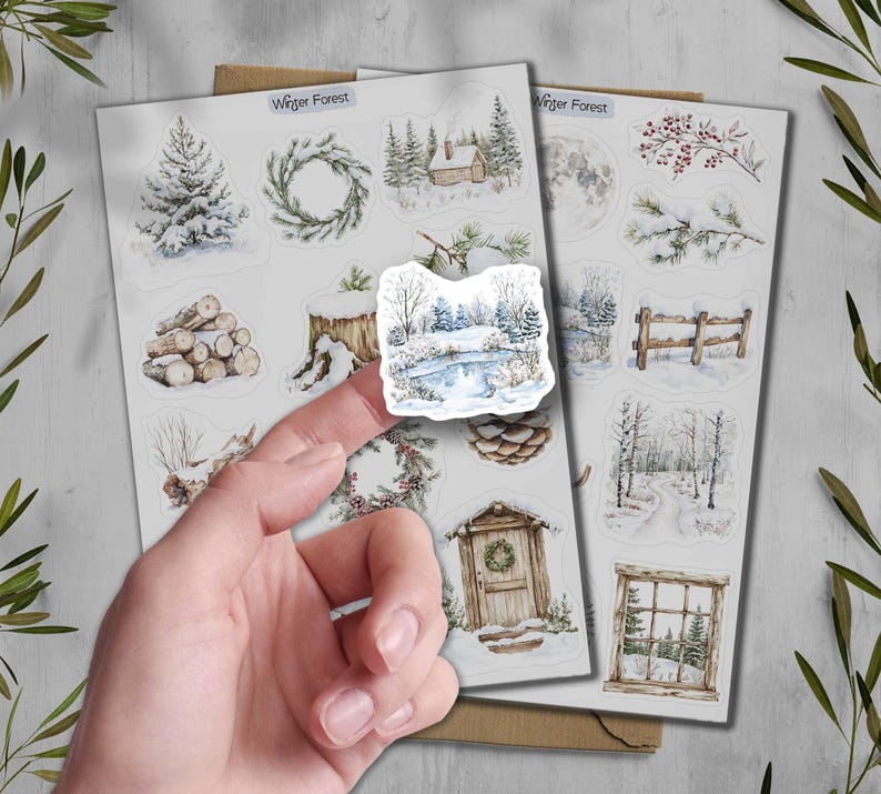 May include: Sheets of winter-themed stickers featuring watercolor illustrations of snowy landscapes, cabins, wreaths, and trees. The text "Winter Forest" is printed at the top of the sheets. The stickers are held by a person's finger.