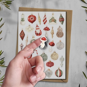 Puede incluir: Una hoja de pegatinas de adornos navide&ntilde;os con varios dise&ntilde;os, incluyendo un mu&ntilde;eco de nieve, Pap&aacute; Noel y setas. Un dedo sostiene una pegatina de seta. Las pegatinas est&aacute;n sobre un fondo blanco con un sobre marr&oacute;n.