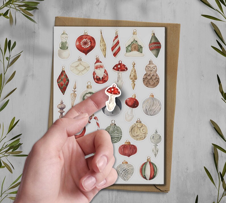 Puede incluir: Una hoja de pegatinas de adornos navide&ntilde;os con varios dise&ntilde;os, incluyendo un mu&ntilde;eco de nieve, Pap&aacute; Noel y setas. Un dedo sostiene una pegatina de seta. Las pegatinas est&aacute;n sobre un fondo blanco con un sobre marr&oacute;n.