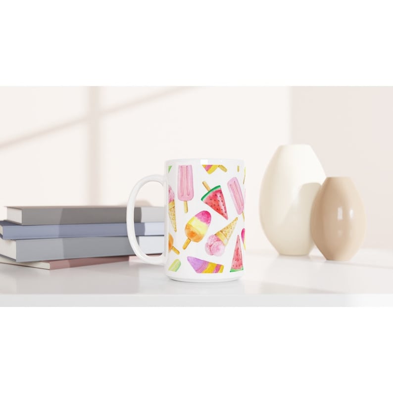 May include: White ceramic mug with a colorful pattern of ice cream cones and popsicles. The ice cream is in shades of pink, yellow, and blue. The popsicles are pink and yellow. The background is white.