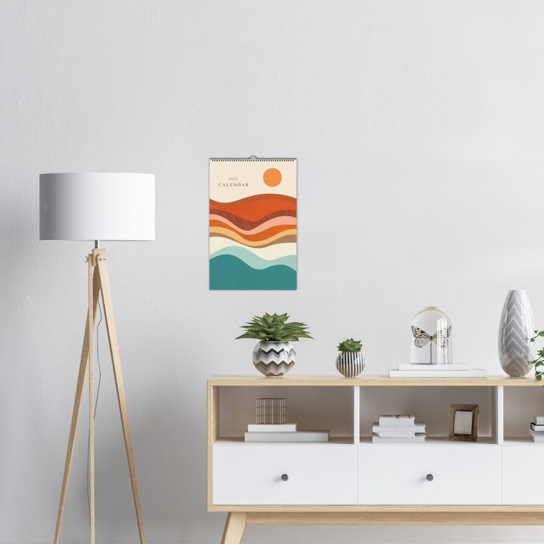 May include: A 2023 calendar with a colourful abstract design featuring a sun, hills, and waves in shades of orange, red, yellow, blue, and green. The calendar is hanging on a white wall above a wooden shelf with decorative items.
