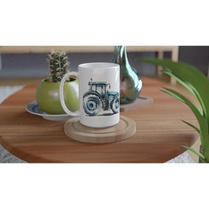 K&ouml;nnte beinhalten: Eine wei&szlig;e Keramiktasse mit einer blauen Aquarell-Illustration eines Traktors auf der Seite. Die Tasse steht auf einem Untersetzer aus Holz.