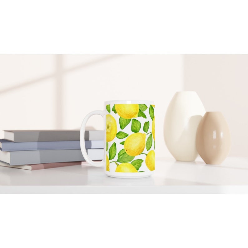K&ouml;nnte beinhalten: Wei&szlig;e Keramiktasse mit Zitronenmotiv. Die Tasse zeigt gelbe Zitronen mit gr&uuml;nen Bl&auml;ttern auf wei&szlig;em Hintergrund.