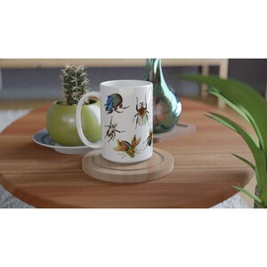 Puede incluir: Taza de cer&aacute;mica blanca con un dise&ntilde;o de escarabajos coloridos. La taza tiene un asa y est&aacute; sobre un posavasos de madera.