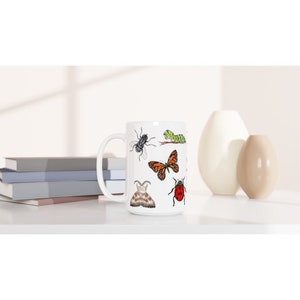 K&ouml;nnte beinhalten: Wei&szlig;e Keramiktasse mit einem bunten Design aus verschiedenen Insekten, darunter ein Schmetterling, ein Marienk&auml;fer, eine Raupe und eine Fliege.