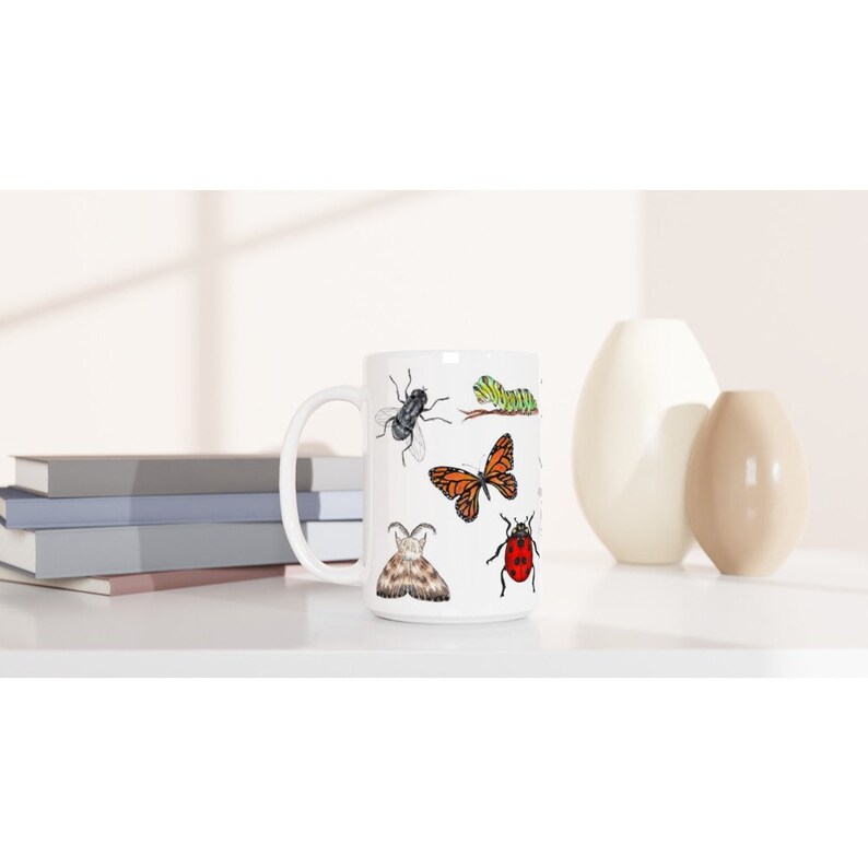 K&ouml;nnte beinhalten: Wei&szlig;e Keramiktasse mit einem bunten Design aus verschiedenen Insekten, darunter ein Schmetterling, ein Marienk&auml;fer, eine Raupe und eine Fliege.