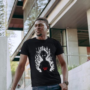 Peut inclure: T-shirt noir avec un motif blanc d'une silhouette dans les flammes et le texte rouge "Mob Psycho 100". Le t-shirt est porté avec un jean bleu et une montre.