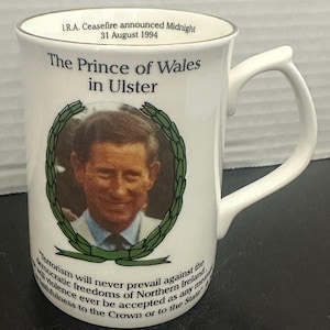 Può includere: Una tazza in ceramica bianca con un bordo dorato. La tazza presenta un'immagine a colori del principe Carlo con il testo "The Prince of Wales in Ulster" e "Terrorism will never prevail against the democratic freedoms of Northern Ireland, and will violence ever be accepted as any means of faithfulness to the Crown or to the State."