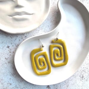 May include: Two yellow, square spiral earrings with silver hooks, presented on a white, organically shaped ceramic plate. The earrings have a glossy finish and a retro design. A white ceramic face is in the background.