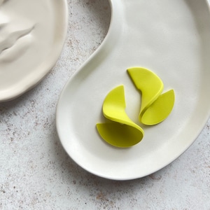 May include: A close-up shot of two white ceramic plates. One plate is a simple oval shape, and the other is a kidney bean shape. Four lime green, curved, petal-like objects are arranged on the kidney bean-shaped plate.