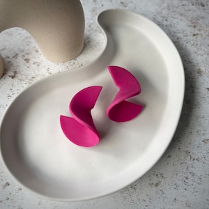 May include: A pair of pink, twisted, geometric earrings. The earrings are made of a smooth, glossy material and have a modern, abstract design.
