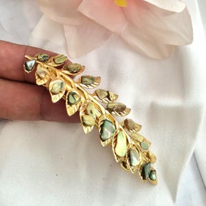 Könnte beinhalten: Goldfarbene Haarspange im Blätterdesign, verziert mit schillernden, oval geformten Akzenten. Die Spange hat einen Spangenstil und befindet sich vor einem weißen Hintergrund mit einer rosa Blume. Die Spange ist etwa 10 cm lang.