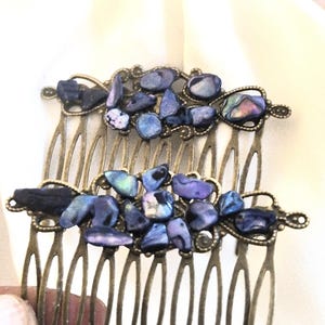 Op de afbeelding: Twee antieke bronzen haarspelden, elk versierd met clusters van blauwe en paarse iriserende stenen. De stenen zijn onregelmatig van vorm en geplaatst tegen een sierlijk filigraan ontwerp. De kammen hebben meerdere tanden.