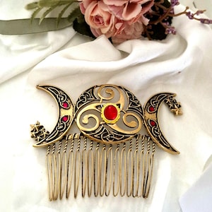 Peut inclure: Un peigne à cheveux doré orné d'un motif triskèle central avec une pierre ovale rouge, flanqué de deux croissants de lune décorés de petites étoiles et d'accents rouges. Le métal présente des motifs de nœuds complexes sur toute la pièce.