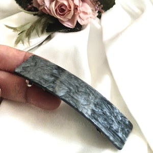 May include: A rectangular hair barrette with a marbled grey and white pattern. The barrette has a silver-coloured clasp. The background includes white fabric and pink flowers.