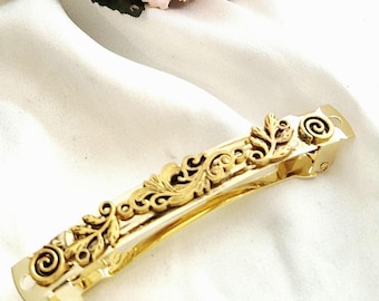 May include: A gold-colored hair clip with an ornate, floral design. The clip features a rectangular shape with detailed scrollwork and leaf patterns. The ends of the clip have a spiral design.