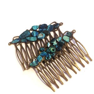 May include: Two hair combs featuring an antique bronze-toned metal base with multiple long, curved teeth. The top of each comb is decorated with an arrangement of iridescent blue and green abalone shell fragments in various irregular shapes.
