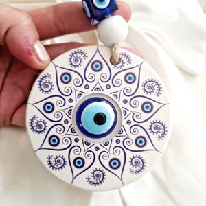 May include: White ceramic round wall hanging with a blue evil eye design. The evil eye is surrounded by a swirling pattern of blue and white. The wall hanging is attached to a white bead and blue evil eye bead string.