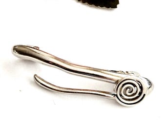 May include: A silver-toned metal hair clip with a spiral design at one end. The clip has a curved shape and a smooth, polished finish. The background includes pink flowers.