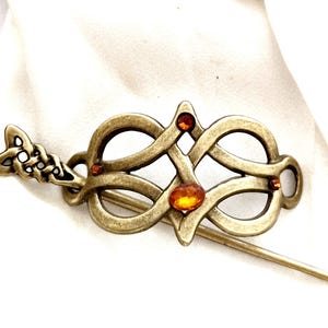 Peut inclure: Une broche en forme de nœud celtique de couleur or avec des pierres en verre de couleur ambre. La broche a une épingle au dos pour l'attacher aux vêtements.