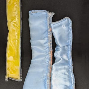 May include: A yellow ice pop in a clear plastic wrapper next to two light blue satin doll boots. The boots have a fluffy trim and a pink accent. The items are on a black background.