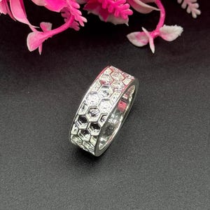Peut inclure: Une bague en argent avec un motif en nid d'abeille. La bague est posée sur un fond noir, avec des fleurs roses en arrière-plan. Le motif de la bague est un motif géométrique répétitif.