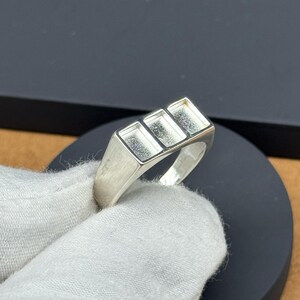 Op de afbeelding: Een zilveren ring met een rechthoekige voorkant met drie rechthoekige inzetstukken. De ring wordt door een gehandschoende hand tegen een donkere achtergrond gehouden. Het ontwerp van de ring is eenvoudig en modern, geschikt voor dagelijks gebruik.