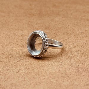 May include: A silver-coloured metal ring blank featuring a circular setting with a decorative rope-style border. The ring has a smooth, plain band and an empty, recessed centre designed to hold a stone or cabochon, showcasing a metallic finish.