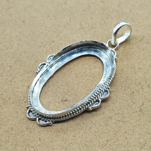 May include: A silver-toned oval pendant setting featuring a hollow centre for a gemstone. The frame is decorated with a twisted rope texture along the outer edge and small circular accents. A sturdy bail is attached to the top for hanging.