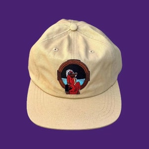 Puede incluir: Gorra de béisbol beige con visera curva y botón en la parte superior. La gorra presenta un parche bordado con una figura con una túnica roja, sobre un fondo azul y marrón. La gorra está sobre un fondo morado.