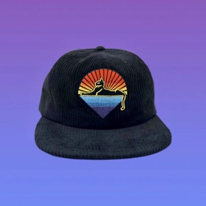 May include: Black corduroy baseball cap with a curved brim. The cap features an embroidered design of a cat silhouette lounging in front of a sunset with orange, yellow, and blue hues. The cap has a rounded crown and a button on top.