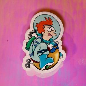 Puede incluir: Una pegatina de dibujos animados de un personaje pelirrojo que lleva un traje espacial azul y un casco, montando un cohete de juguete amarillo con una hélice azul y verde.