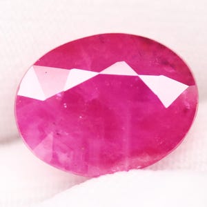 May include: Oval-shaped, faceted ruby gemstone. The gem is a deep, saturated pinkish-red color with a polished surface. The facets create a play of light and shadow, enhancing the stone's brilliance. The ruby is set against a white background.