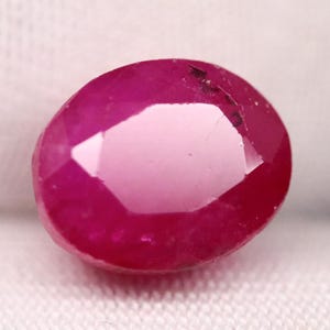 May include: A faceted, oval-shaped ruby gemstone. The gem is a deep, saturated red color with a polished surface. The ruby is set against a white background.