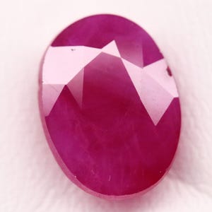 May include: A faceted, oval-shaped ruby gemstone. The gem is a deep, saturated red color with multiple facets that reflect light. The gemstone is set against a white background.