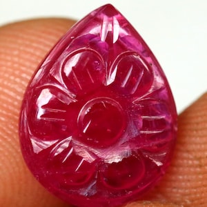 May include: A teardrop-shaped, carved ruby gemstone. The deep red gem features a floral design carved into its surface. The gemstone is held between two fingers, showcasing its size and detail. The carving is intricate and well-defined.