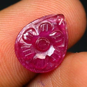Può includere: Una gemma di rubino intagliata a forma di goccia. La gemma rosso intenso presenta un disegno floreale intagliato sulla sua superficie. L'intaglio è dettagliato, con petali visibili e un elemento circolare centrale. La gemma è tenuta tra due dita.