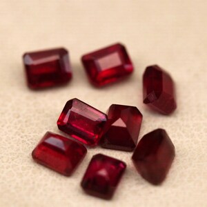 May include: A collection of faceted, rectangular, and irregularly shaped red gemstones. The gems are scattered on a light beige surface, showcasing their deep red color and reflective facets. The gemstones vary in size and shape, suggesting a collection of loose stones.