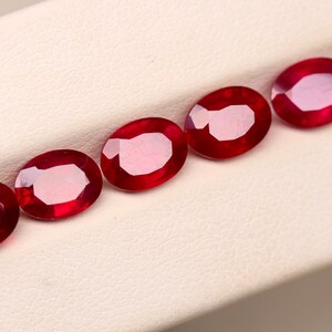 May include: Five oval-shaped, deep red gemstones are arranged diagonally across a white surface. The gems have a polished, reflective surface, and a lighter, pinkish interior. These rubies are likely for jewelry making.