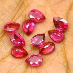 Puede incluir: Una colección de diez gemas facetadas en forma de pera, de color rosa intenso. Las gemas están dispuestas sobre una superficie marrón claro, mostrando su talla brillante y su color vibrante. Cada piedra refleja la luz.