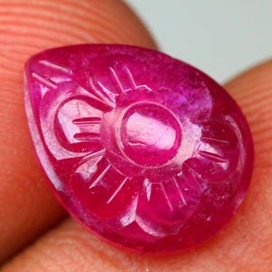 Könnte beinhalten: Ein tropfenförmiger, geschnitzter Rubin-Edelstein. Der tiefrosa Stein weist ein Blumenmuster mit einem zentralen Kreis und radialen Linien auf. Der Edelstein wird zwischen zwei Fingern gehalten, wodurch seine polierte Oberfläche und die komplizierten Details hervorgehoben werden.