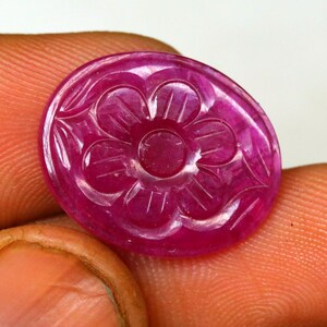 May include: A round, polished ruby gemstone with a carved floral design. The gemstone is a deep pink color and is held between fingers. The flower carving has detailed petals and a central circle.
