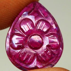 May include: A carved, teardrop-shaped gemstone in shades of pink and red. The gemstone features a floral design carved into its surface. The carving is detailed and intricate, with visible lines and curves. The gemstone is held between two fingers.