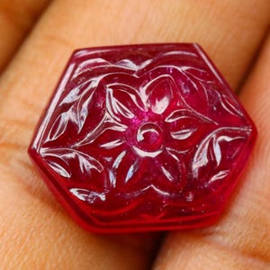 May include: A faceted, hexagonal ruby gemstone with a floral design carved into its surface. The deep red stone has a polished finish, and the carved details are highlighted by the light. The gemstone is held in a hand.