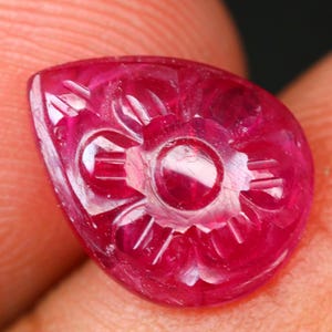 May include: A carved ruby gemstone, teardrop-shaped. The deep red gemstone features a floral design carved into its surface. The carving includes a central circle with radiating lines and petal-like shapes. The ruby is held between two fingers.