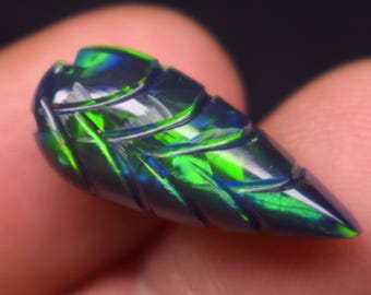 May include: A carved black opal gemstone featuring a leaf-like shape with deep grooves and vibrant flashes of neon green and blue light. The surface of the stone is polished and reflective, showcasing a play of color across its dark, iridescent body.
