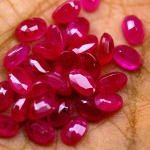 May include: A collection of small, oval-shaped, faceted rubies with a vibrant, deep pinkish-red hue. Each gemstone features a polished surface with multiple geometric facets that reflect light, creating a sparkling appearance across the stones.