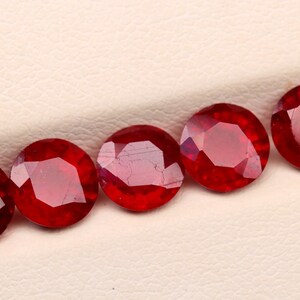 May include: Five round, faceted, deep red gemstones are arranged in a diagonal line. The gems have a brilliant cut, reflecting light and showcasing their rich color. The background is a neutral, off-white surface, highlighting the gemstones.